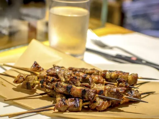 Arrosticini, uno dei piatti tipici abruzzesi, serviti su carta paglia con un bicchiere di vino bianco sullo sfondo.