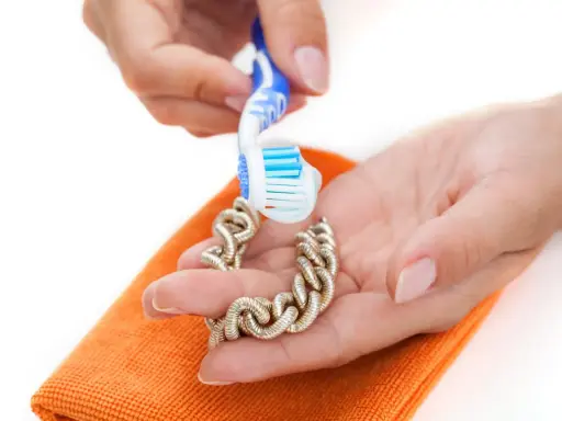 Manos limpian una pulsera de oro con un cepillo de dientes y un paño naranja. Aprende como limpiar el oro para que brille.