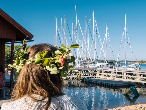 En flicka med blomsterkrans ser ut över en hamn med segelbåtar. En perfekt dag för en resa till Åland.