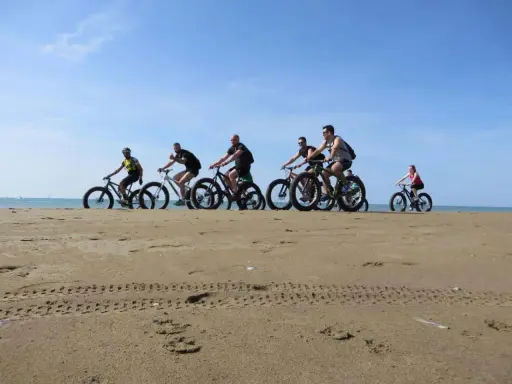 Gruppo di ciclisti su fat bike lungo la ciclabile di Caorle, sulla sabbia dorata con il mare sullo sfondo.