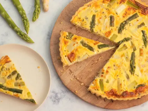 Herzhafte Spargel Quiche mit frischem Spargel, goldbraun gebacken. Ein Stück auf einem Teller, der Rest auf einem Holzbrett.