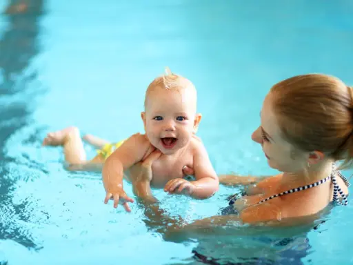 Glückliches 8 Wochen altes Baby mit Mutter im Schwimmbad. Das Baby lacht und planscht im Wasser, während die Mutter es festhält.