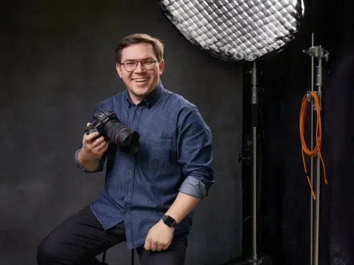 Uśmiechnięty mężczyzna z aparatem, gotowy poprowadzić kurs fotografii dla początkujących. W tle sprzęt studyjny.