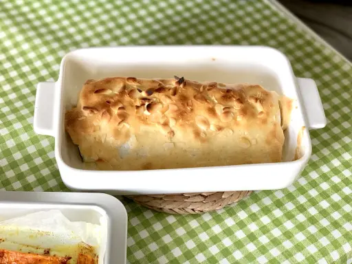 Bacalao gratinado con alioli, cubierto de almendras tostadas, en una fuente blanca sobre mantel de cuadros verdes.