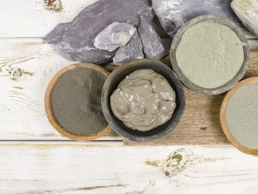 Pots de différentes argiles, dont une argile verte pour dégonfler, sur un fond en bois blanc et brut.