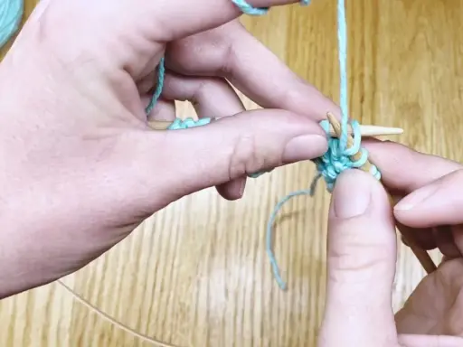Manos tejiendo punto ojo de perdiz con hilo turquesa y agujas de madera.