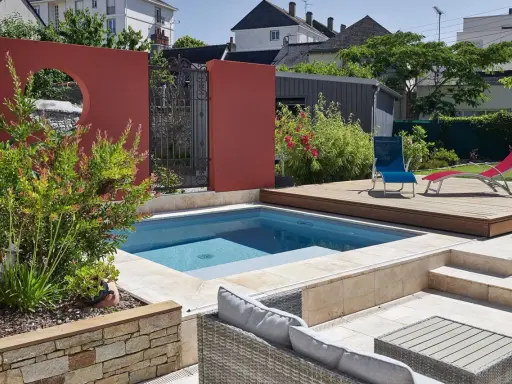 Une piscine moderne avec une belle décoration autour piscine : mur rouge, terrasse en bois, mobilier de jardin et végétation luxuriante.