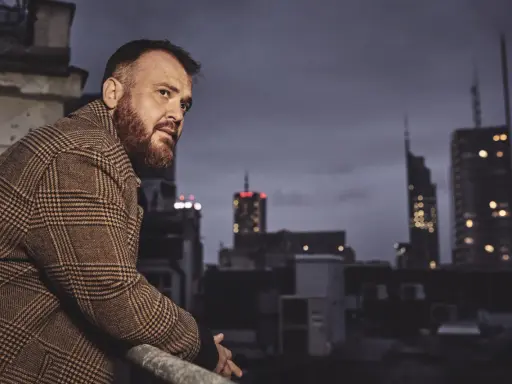 Chmielarz długa noc. Mężczyzna w płaszczu w kratę spogląda na nocne miasto z dachu.
