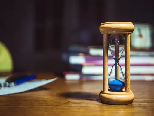 Clessidra con sabbia blu su una scrivania, a simboleggiare quante ore devono passare tra un turno e l'altro. Libri e fogli sullo sfondo.