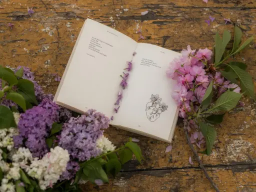 Un libro aperto con poesie d'amore passionali anonime, decorato con fiori lilla e rosa su un tavolo di legno consumato.