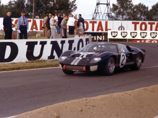 Ein schwarzer Ford GT40 Mk3 mit der Startnummer 2 rast auf der Rennstrecke, während Zuschauer und Werbetafeln im Hintergrund zu sehen sind.