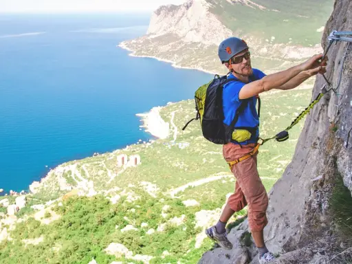 Mężczyzna na via ferrata Krucze Skały, zmagający się ze stromą ścianą skalną, z widokiem na błękitne morze i zielone wybrzeże.