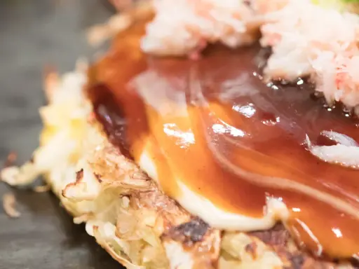 Nahaufnahme eines Okonomiyaki mit cremiger Mayonnaise und würziger Okonomiyaki-Sauce, garniert mit zarten Krabbenfleischstückchen.
