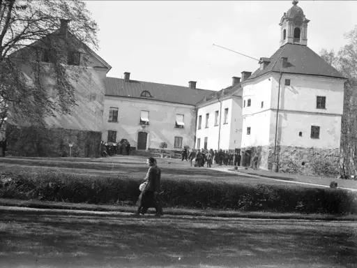 En grupp människor samlas framför Rydboholms slott. En kvinna går över gräsmattan.