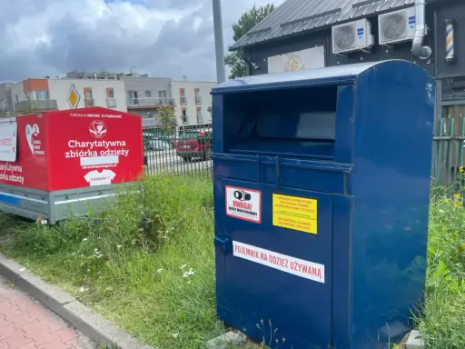 Kontener na odzież używaną obok czerwonego przyczepy z napisem "Charytatywna zbiórka odzieży". To miejsce, gdzie można oddać tekstylia, odpady ubraniowe, zamiast wyrzucać.