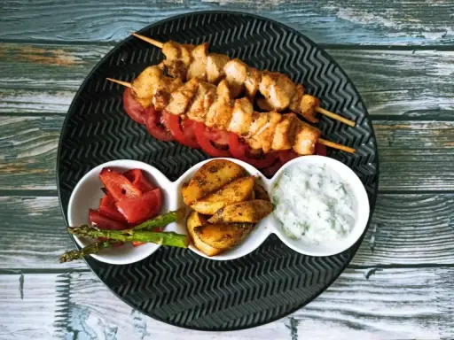 Dwa szaszłyki souvlaki z kurczaka na talerzu z pomidorami, ziemniakami, szparagami i sosem.