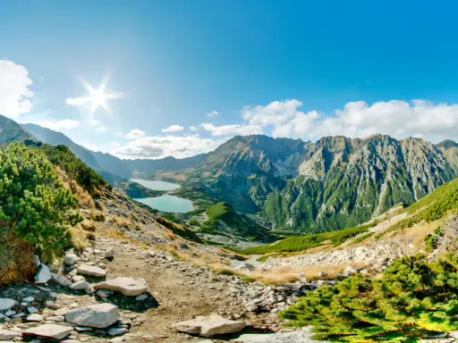 Słoneczna panorama Tatr z dwoma jeziorami w dolinach. Kamienista ścieżka prowadzi przez zielone zbocza porośnięte kosodrzewiną.