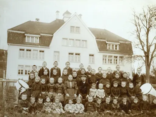Schulkinder und Lehrer posieren vor einem Gebäude. Die Kleidung deutet auf eine Zeit hin, in der Armut, also Pauperismus, ein großes Thema war.