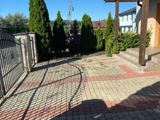 Nowy pomysł na stary chodnik: kostka brukowa w odcieniach beżu i czerwieni tworzy ciekawy wzór przed domem, otoczonym zielenią i ogrodzeniem.