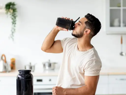 Mann trinkt Proteinshake aus schwarzem Shaker. Daneben steht ein großer Behälter mit Pulver. Wozu Protein**pulver**? Für Muskelaufbau und Erholung.