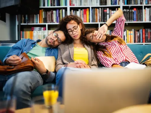 Drei erschöpfte Studenten schlafen auf einer Couch, umgeben von Büchern. Der abi deutsch-Test war wohl zu anstrengend.