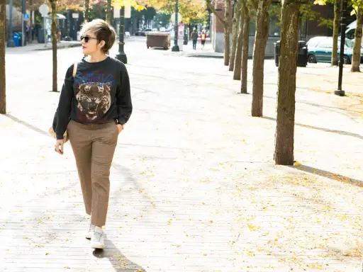 A woman walks down a tree-lined street in a trendy plaid pants outfit, featuring a graphic sweatshirt and white sneakers.