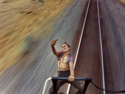 Mike Brodie, un giovane ribelle, fa il gesto del dito medio mentre viaggia su un treno merci, sfidando il mondo con uno sguardo provocatorio.