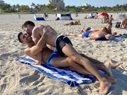 Two men embrace on a towel at Haulover Beach, a popular spot for gay beachgoers. Other sunbathers relax on the sand in the background.