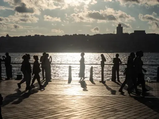 Ludzie spacerują i łowią ryby na promenadzie nad morzem. Słońce odbija się w wodzie, tworząc złoty blask.