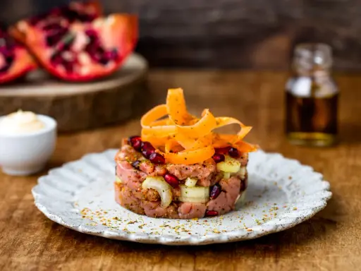 Deliziosa tartare di manzo con melograno, cipolla e carote a spirale, servita su un piatto rustico.