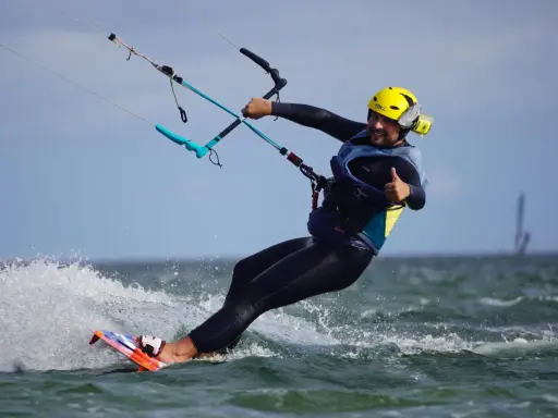 Uśmiechnięty mężczyzna w żółtym kasku i piance cieszy się jazdą na desce. To świetna nauka kitesurfingu!