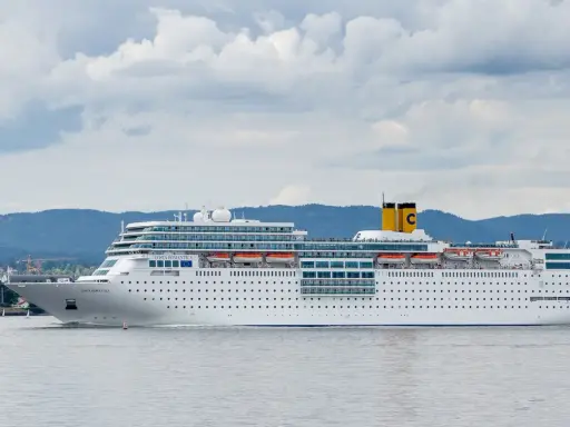 La nave da crociera Costa Romantica naviga in acque calme, con montagne verdi sullo sfondo e un cielo nuvoloso.