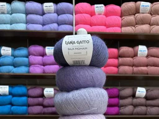 En stapel med tre härvor av lyxigt mohair silke garn i olika lila nyanser. Bakom syns hyllor fyllda med garn i många färger.