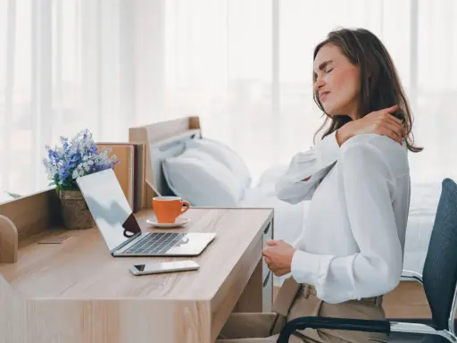 Femme se massant l'épaule, assise à son bureau. La mauvaise position bureau peut causer des douleurs.