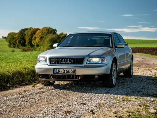 Silberner Audi A8 D2 W12 auf Schotterweg, umgeben von grünen Feldern und Bäumen im Herbstlicht.
