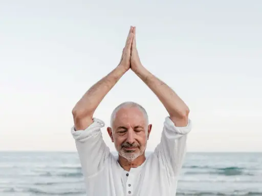 Mężczyzna z zamkniętymi oczami, ręce złożone nad głową, praktykuje mindfulness co to. Spokój nad morzem.