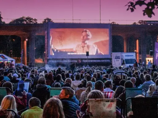 Stor publik njuter av utomhusbio i Stockholm under en rosa kvällshimmel. En stor duk visar en filmscen.