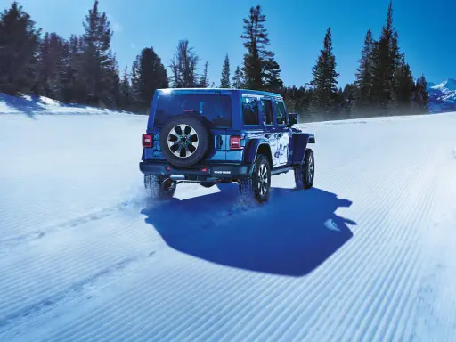 Un blu Jeep Wrangler affronta la neve con pneumatici Toyo Open Country AT3, lasciando tracce su un sentiero battuto tra alberi innevati e montagne.