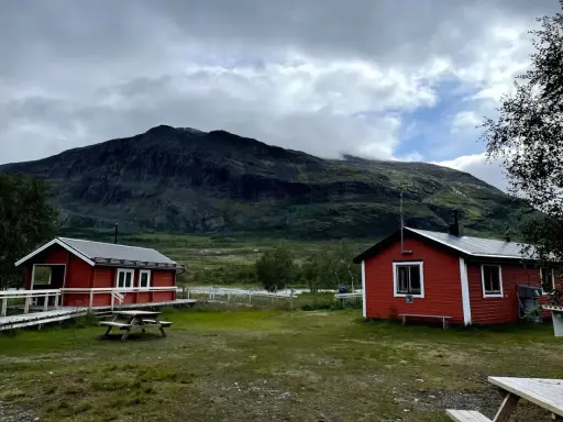 Två röda stugor i Abisko Nikkaluokta med en bergig, molntäckt bakgrund. En picknickbänk står på gräsmattan.