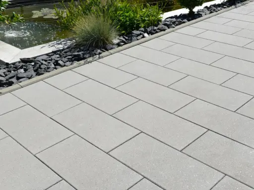 Fertig verlegte Randsteine begrenzen einen modernen Weg aus hellgrauen Platten, daneben ein Teich mit Wasserspiel und Zierkies.