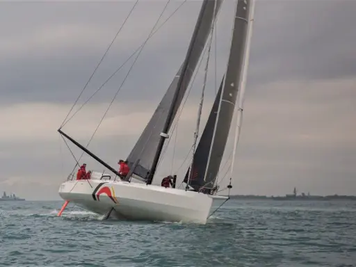 Ein IMOCA 60 segelt auf stürmischer See, die Crew in roten Jacken bereitet sich auf die nächste Welle vor.
