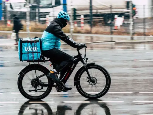 Kurier na rowerze z niebieską torbą Wolt, jadący w deszczu. To przesyłka bez możliwości śledzenia, dostarczana szybko i sprawnie.