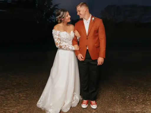 A bride in a lace gown and groom in a rust-colored jacket and black pants, sporting red sneakers, showcase their semi-formal attire for wedding.