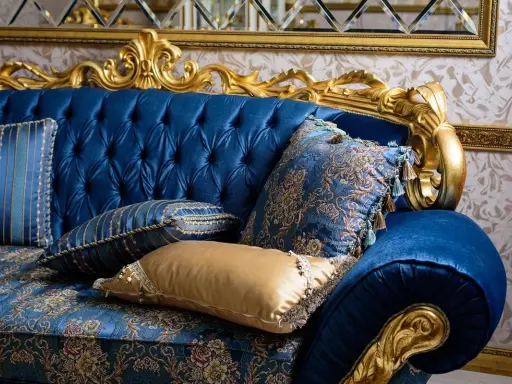 Canapé bleu profond, capitonné, aux ornements dorés, style baroque. Coussins aux motifs floraux et rayés, franges et pompons ajoutent une touche luxueuse.