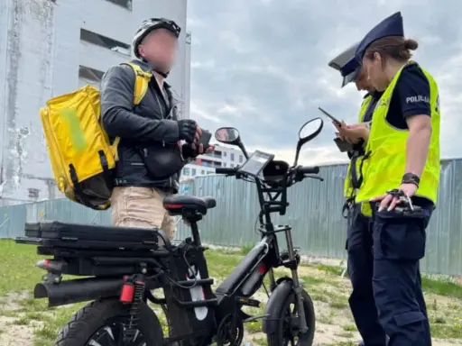 Dostawca z żółtą torbą na plecach rozmawia z policjantkami obok swojego roweru elektrycznego bez pedałowania.