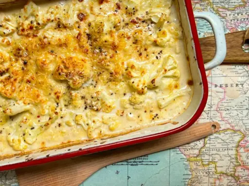 Coliflor gratinado con bechamel cremosa, cubierto de queso dorado y pimienta negra, servido en una fuente roja sobre un mapa antiguo.