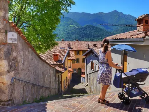 Una donna spinge un passeggino con neonati in montagna, tra case con tetti rossi e un panorama mozzafiato.