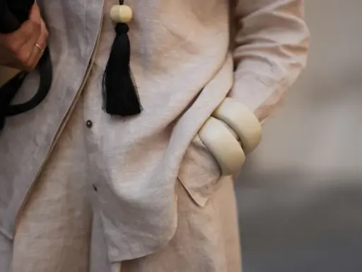 Dettaglio di un outfit in lino materiale, con una mano che tiene una borsa nera, un ciondolo con nappa nera e bracciali color crema.