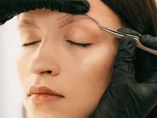 Une main gantée retire délicatement un poil incarné au sourcil d'une femme aux yeux fermés.