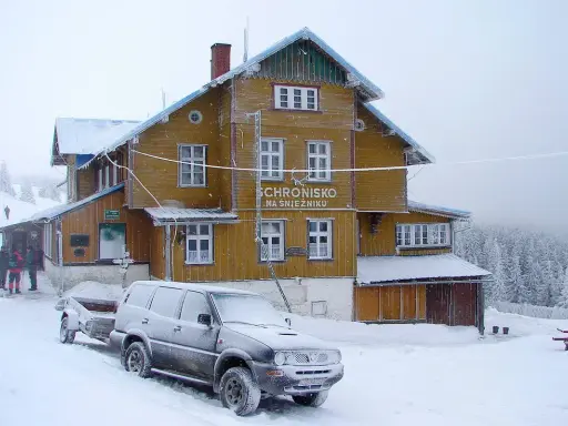 Schronisko na Śnieżniku w zimowej scenerii. Samochód z przyczepą i turyści przy wejściu. Masyw Śnieżnika i jego szlaki w śniegu.